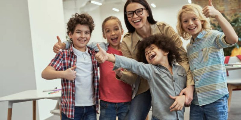 We are the best. Happy school team of four cute little pupils and their friendly female teacher posing together, standing in the classroom. Childhood and education concept