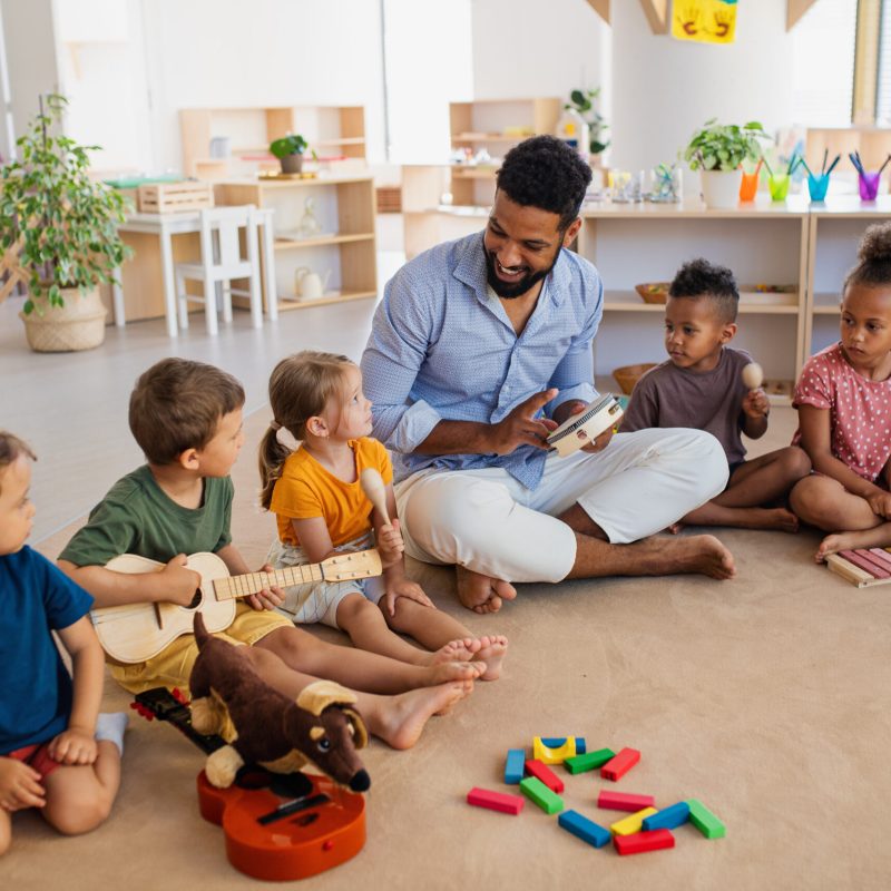 A group of small nursery school children with man teacher sitting on floor indoors in classroom, playing musical instruments.