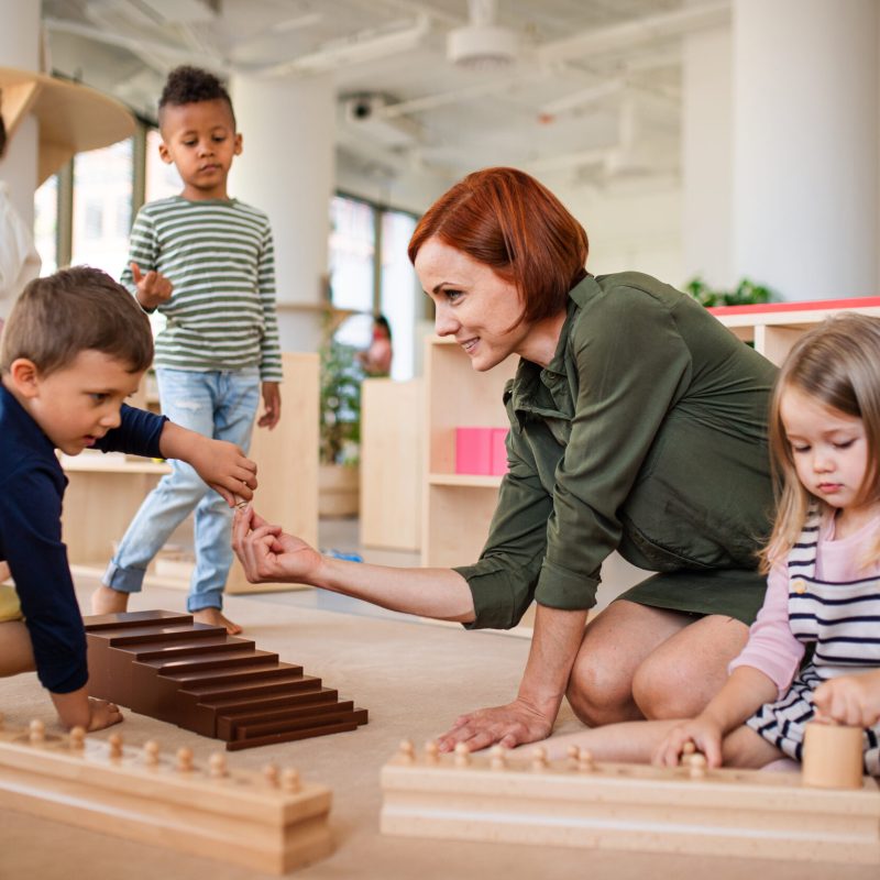 Group of small nursery school children with teacher sitting on floor indoors in classroom, montessori learning concept.
